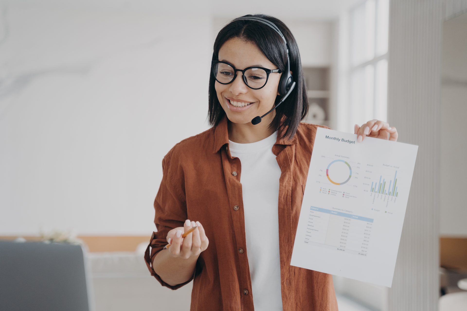 Smiling female manager in headset shows document with statistics while online conference at laptop